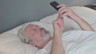 Yorgun ihtiyar yatakta akıllı telefon kullanıyor.