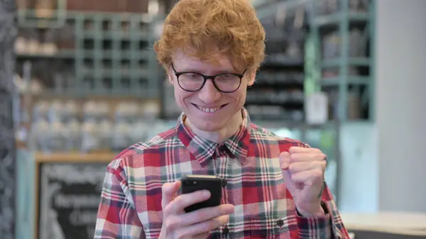 Портрет молодого рыжего молодого человека, празднующего успех на смартфоне — стоковое фото