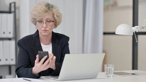 Old Businesswoman Navigation sur Internet sur Smartphone tout en travaillant sur ordinateur portable — Photo