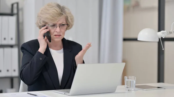 Vieille femme d'affaires en colère sur Smartphone tout en travaillant sur ordinateur portable — Photo