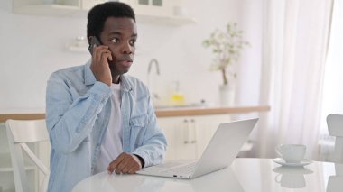 Bilgisayarlı Afrikalı adam evde telefonla konuşuyor.