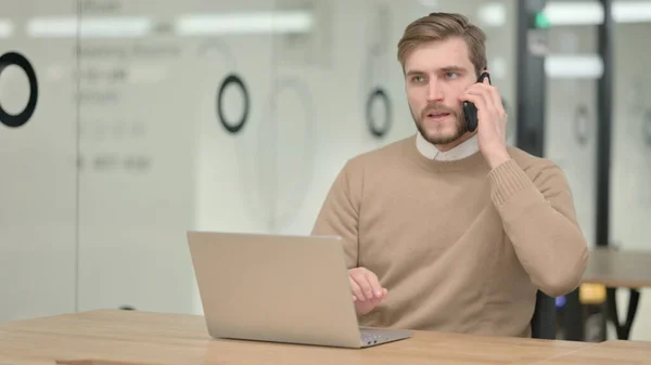 Молодой человек разговаривает по телефону во время работы над ноутбуком — стоковое фото