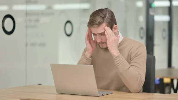 Hombre joven con portátil con dolor de cabeza en la oficina —  Fotos de Stock