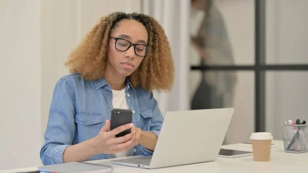 Africká žena pomocí chytrého telefonu a notebooku v kanceláři — Stock fotografie