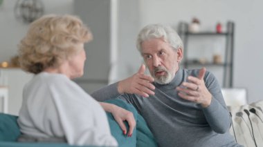 Yaşlı adam yaşlı kadınla konuşuyor, koltukta oturuyor.