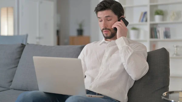 Jongeman met laptop thuis telefonisch aan het praten — Stockfoto