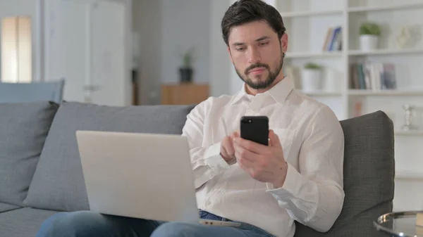 Uomo concentrato che lavora su smartphone e laptop a casa — Foto Stock