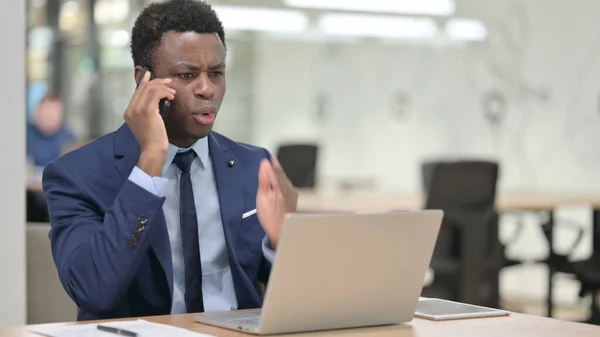 愤怒的非洲商人一边用智能手机一边用笔记本电脑讲话 — 图库照片
