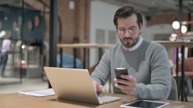 Ofiste dizüstü bilgisayar kullanırken akıllı telefon kullanan genç adam. 