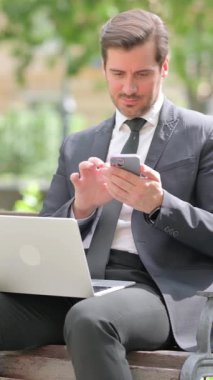 Orta yaşlı iş adamı telefonda konuşuyor ve dışarıda dizüstü bilgisayar kullanıyor.