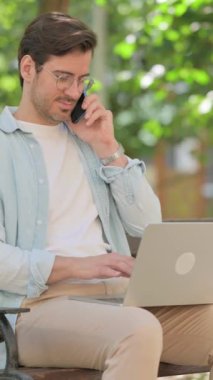 Sıradan genç adam telefonda konuşuyor ve dışarıda dizüstü bilgisayar kullanıyor.