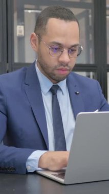 Dikey, Takım Elbiseli Adam Ofisteki Dizüstü bilgisayarda çalışırken Kameraya Bakıyor