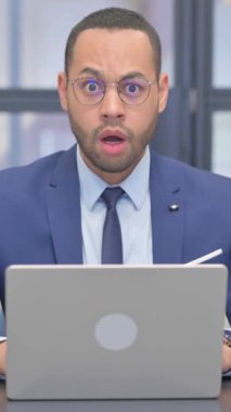 Dikey, Şok Edilmiş Karışık Irk İşadamı Bilgisayarda çalışırken Kameraya Bakıyor