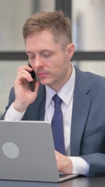 Dikey video, Genç İşadamı Laptop 'ta çalışırken telefonla konuşuyor