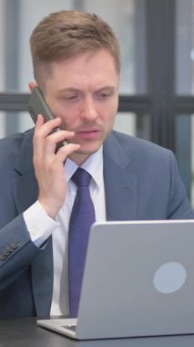 Dikey video, Kızgın İşadamı Laptop 'ta çalışırken telefonda konuşuyor