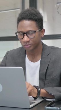 Ofiste dizüstü bilgisayarla çalışırken kameraya bakan dikey, genç Afrikalı adam