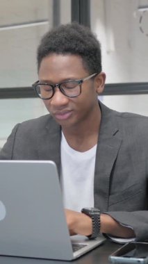 Ofisteki dizüstü bilgisayarda çalışırken kameraya gülümseyen dikey, genç Afrikalı adam