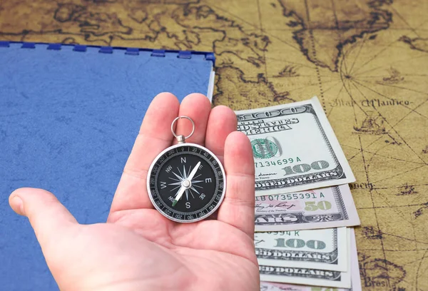 Classic round compass in hand on background of notepad, dollars and old vintage map of world as symbol of tourism with compass, travel with compass and outdoor activities with compass