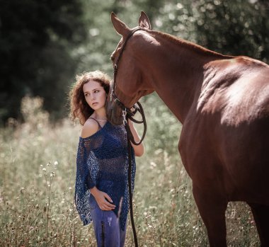 Güzel kıvırcık saçlı bir kız kırmızı bir atın yanında duruyor.