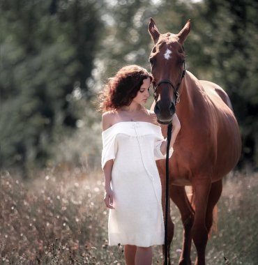 Güzel kıvırcık saçlı bir kız kırmızı bir atın yanında duruyor.