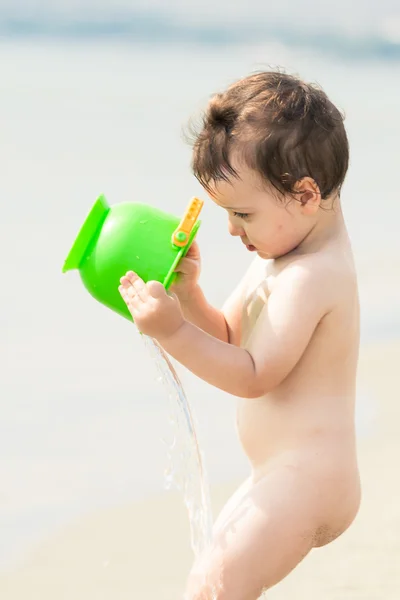 Lindo niño en una playa vertiendo agua en sí mismo con un cubo . — Foto de Stock