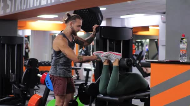 Gym-Unterricht mit einem jungen lustigen Trainer Mann Eine junge Anfängerin schüttelt ihre Beine auf dem Simulator — Stockvideo