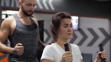 Genç ve komik bir antrenörle beden eğitimi dersi. Acemi bir kadın simülatörde sırtını ve kollarını sallıyor.