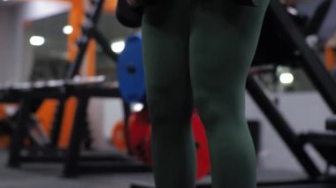 Eğitmen Acemi 'den beden eğitimi dersi. Genç bir kadın, dambıllarıyla sırtını öne doğru sallıyor.