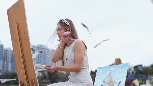 Jonge blonde vrouw schildert beeld op het strand op open lucht tegen de achtergrond van vliegende meeuwen — Stockvideo