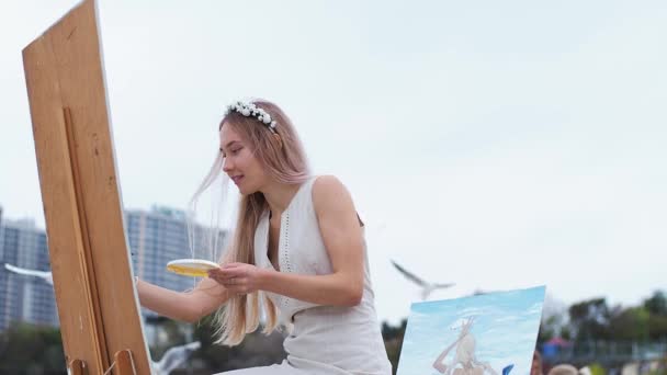 Jovem loira pinta quadro na praia Ao ar livre Contra o fundo de gaivotas voadoras — Vídeo de Stock