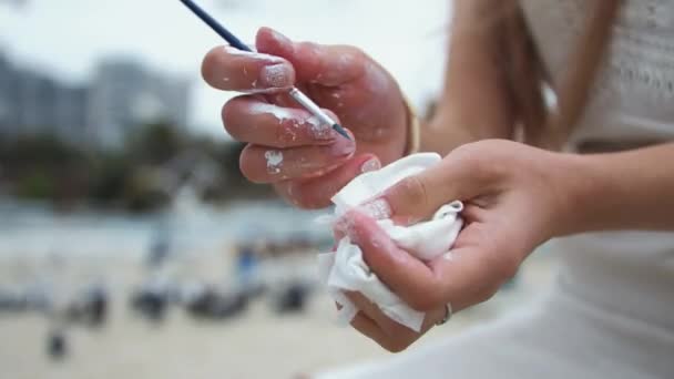 Jovem loira pinta um quadro na praia da cidade. No ar livre Limpa a pintura de mãos — Vídeo de Stock