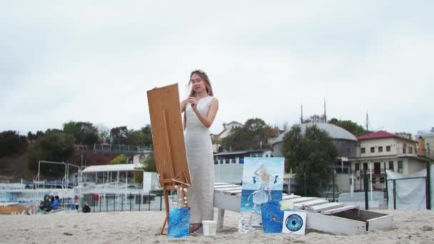 Молодая блондинка рисует картину на пляже под открытым небом на фоне летающих чаек — стоковое видео