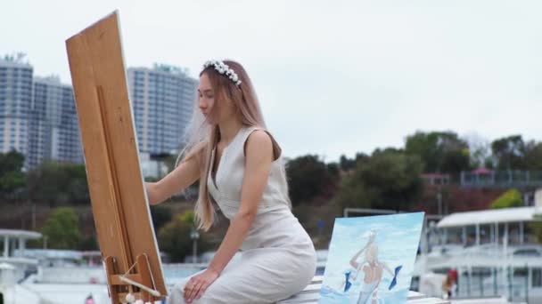 Jonge blonde vrouw schildert beeld op het strand op open lucht tegen de achtergrond van vliegende meeuwen — Stockvideo