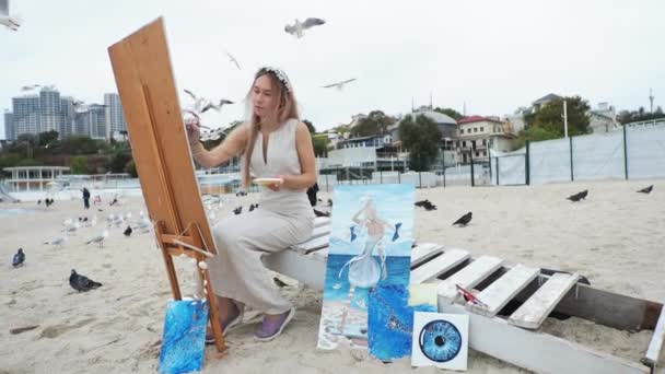 Junge blonde Frau malt Bild am Strand Unter freiem Himmel Vor dem Hintergrund fliegender Möwen — Stockvideo