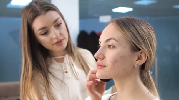 Fiatal barna nő make-up művész fest egy szőke lány egy szépségszalonban Alkalmazza toning krém akne — Stock videók