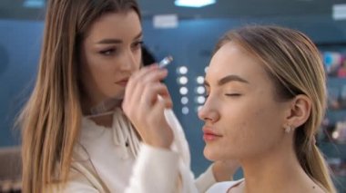 Genç esmer kadın makyaj sanatçısı sarışın bir kızı güzellik salonunda boyuyor kaşlarını boyuyor.