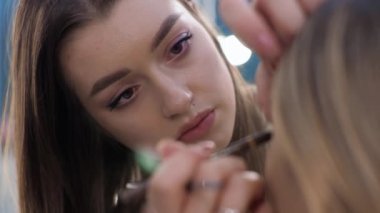 Genç esmer kadın makyaj sanatçısı. Güzellik salonunda sarışın bir kız resmediyor. Göz çevresini boyuyor.