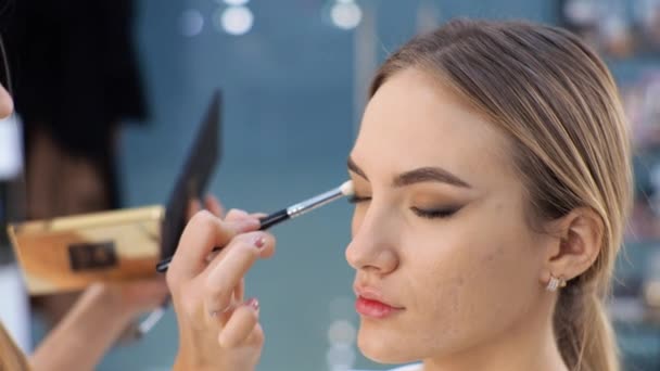 Joven morena maquilladora pinta a una chica rubia en un salón de belleza Eyeshadows — Vídeo de stock