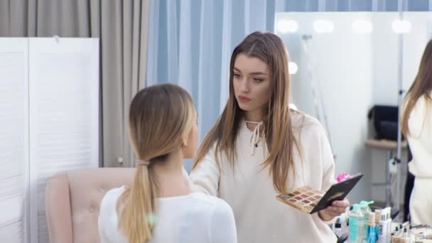 Jonge brunette vrouw make-up kunstenaar schildert een blond meisje in een schoonheidssalon Eyeshadows — Stockvideo