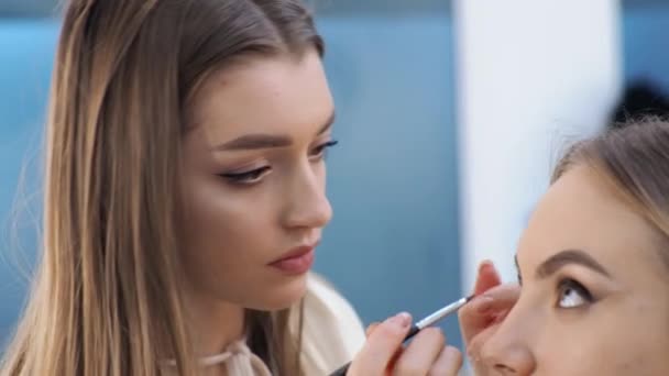 Νεαρή μελαχρινή γυναίκα make-up καλλιτέχνης ζωγραφίζει ένα ξανθό κορίτσι σε ένα σαλόνι ομορφιάς Eyeshadows — Αρχείο Βίντεο