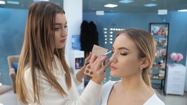 Νεαρή μελαχρινή γυναίκα make-up καλλιτέχνης ζωγραφίζει ένα ξανθό κορίτσι σε ένα σαλόνι ομορφιάς Eyeshadows — Αρχείο Βίντεο