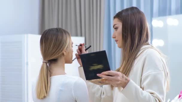 Joven morena maquilladora pinta a una chica rubia en un salón de belleza Eyeshadows — Vídeo de stock