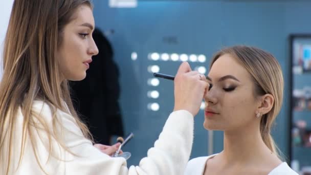 Jonge brunette vrouw make-up kunstenaar schildert een blond meisje in een schoonheidssalon Eyeshadows — Stockvideo