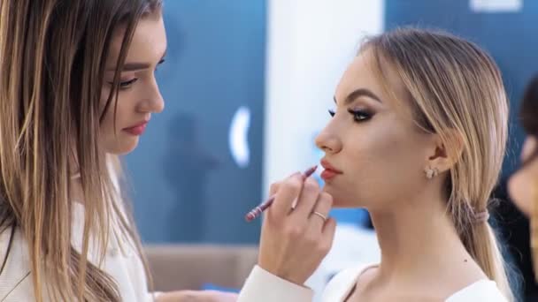 Genç esmer kadın makyaj sanatçısı sarışın bir kızı güzellik salonunda boyuyor dudaklarını boyuyor. — Stok video