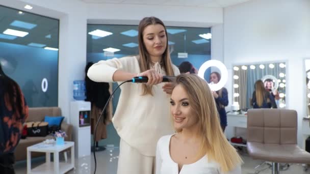 Jovem morena mulher cabeleireiro faz styling no cabelo longo da menina loira — Vídeo de Stock