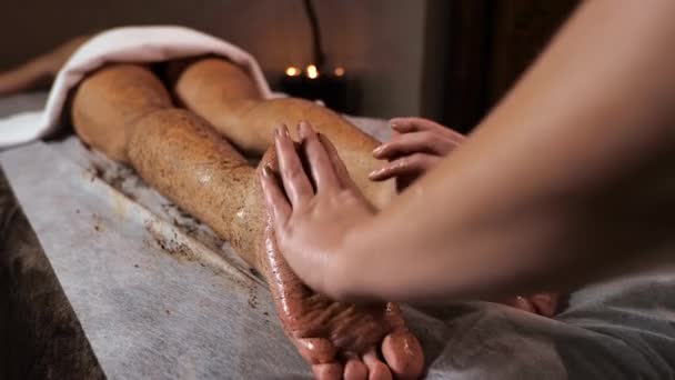 Mulher massagista faz massagem esfregando das pernas massagem bagaço de café em um fundo morno escuro — Vídeo de Stock