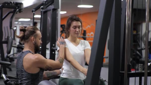 Aula de ginástica com um jovem treinador engraçado Uma mulher iniciante balança as costas e os braços no simulador — Vídeo de Stock