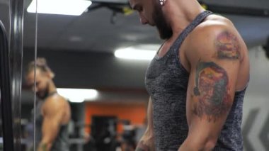Genç adam spor salonunda çalışıyor. Ciddi şekilde odaklanmış. Kolların bisepslerini simülatöre doğru sallıyor.