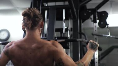 Genç adam spor salonunda çalışıyor. Kollarını sallıyor ve simülatöre sarılıyor. Çubuğu aşağı çekiyor.