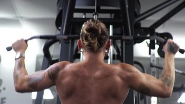 Genç adam spor salonunda çalışıyor. Kollarını sallıyor ve simülatöre sarılıyor. Çubuğu aşağı çekiyor.